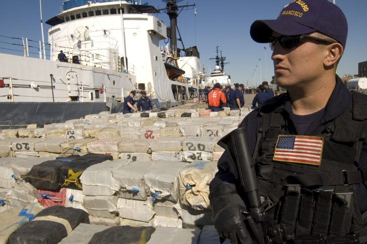US Coast Guard security member standing on the dock next two large stacks of seized drugs and contraband with a large ship in the background.