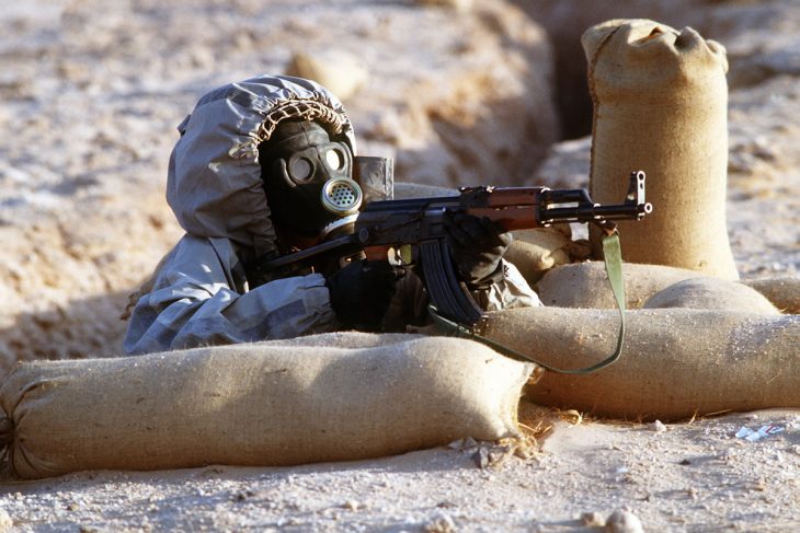 Image of soldier in chemical warfare protective suit and an AK-47 sitting behind sandbags.